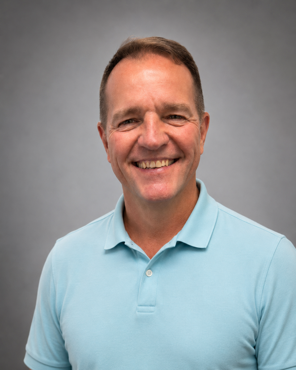 A smiling man with short dark hair, wearing a light blue polo shirt and dark jeans, standing against a plain light-colored wall.