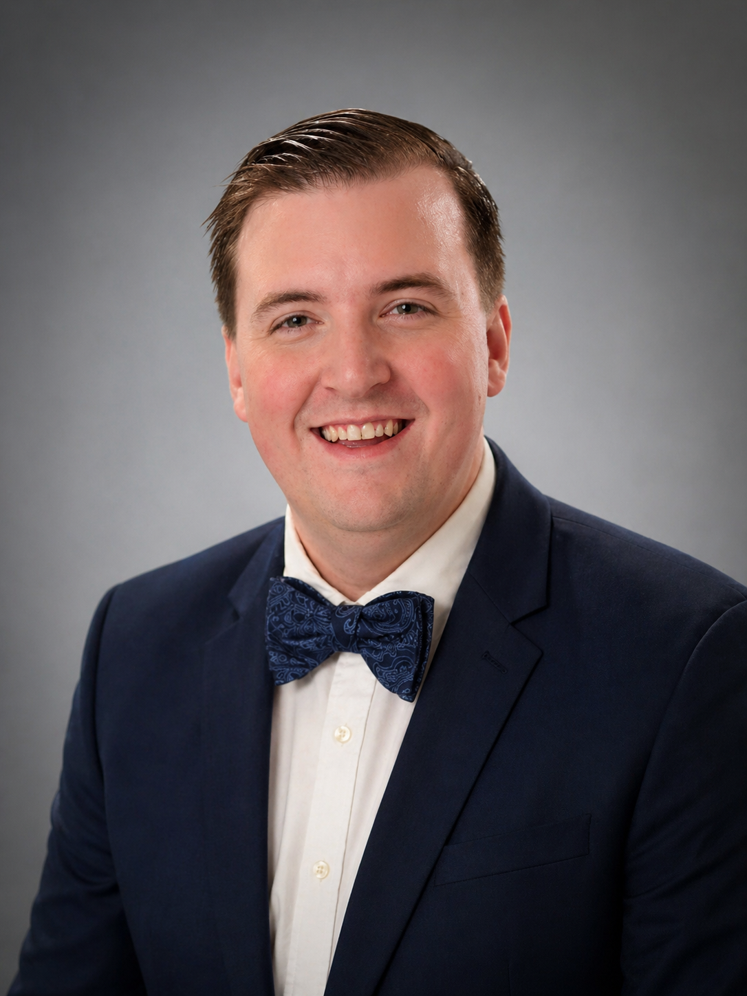 A young man with brown hair, smiling, wearing a navy suit, white dress shirt, and blue patterned bow tie, against a gray background.