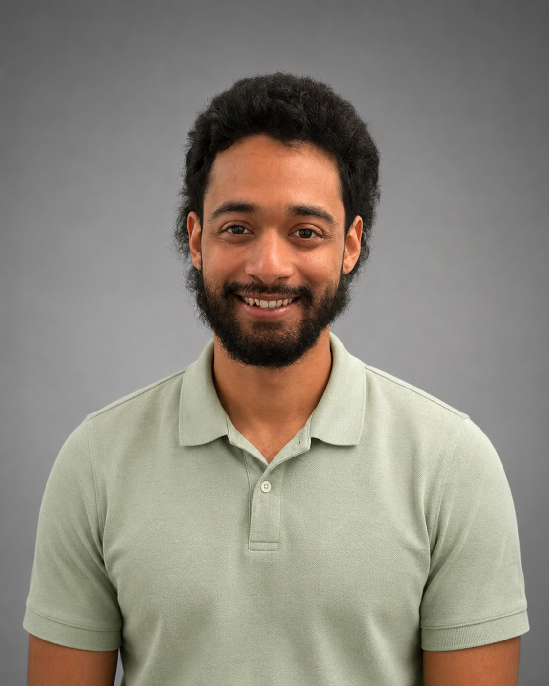 A smiling man with curly hair and a beard, wearing a beige sweater over a collared shirt, standing against a plain off-white background.