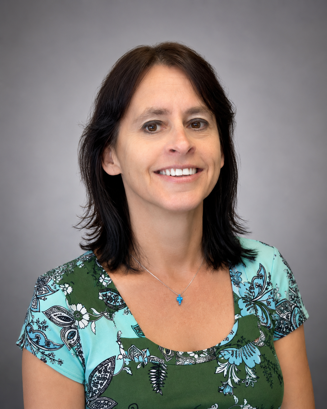 A woman with dark hair and a floral patterned shirt smiling in front of a wooden wall.