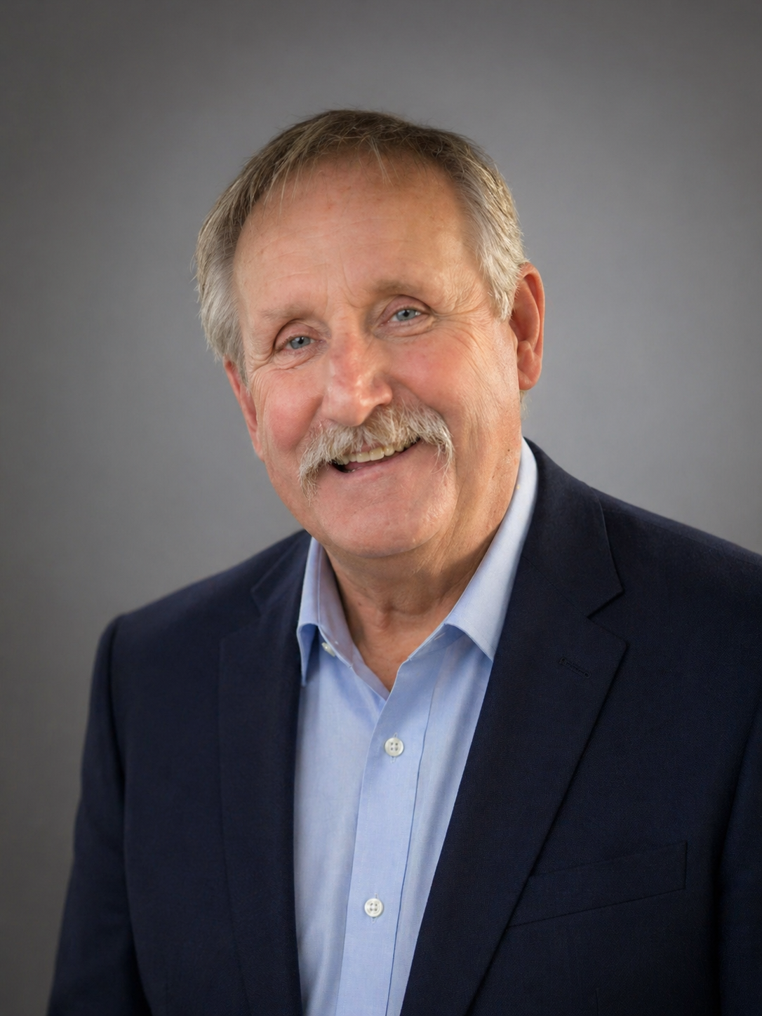 Headshot of a smiling elderly man with light hair and mustache, wearing a dark blazer and light blue shirt, against a gray background.