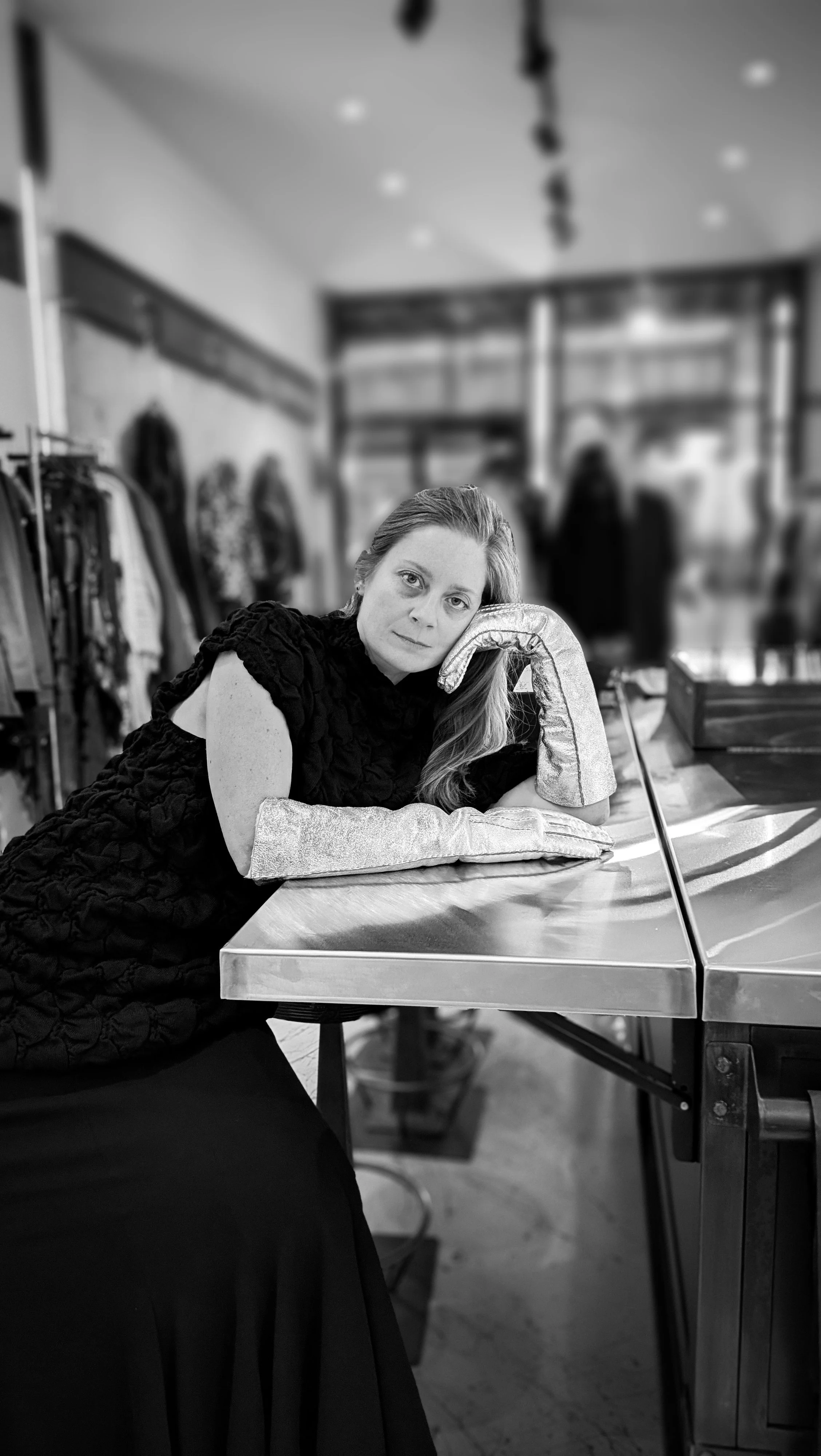 Black-and-white portrait of an artist seated inside an independent fashion boutique, wearing sculptural silver gloves.