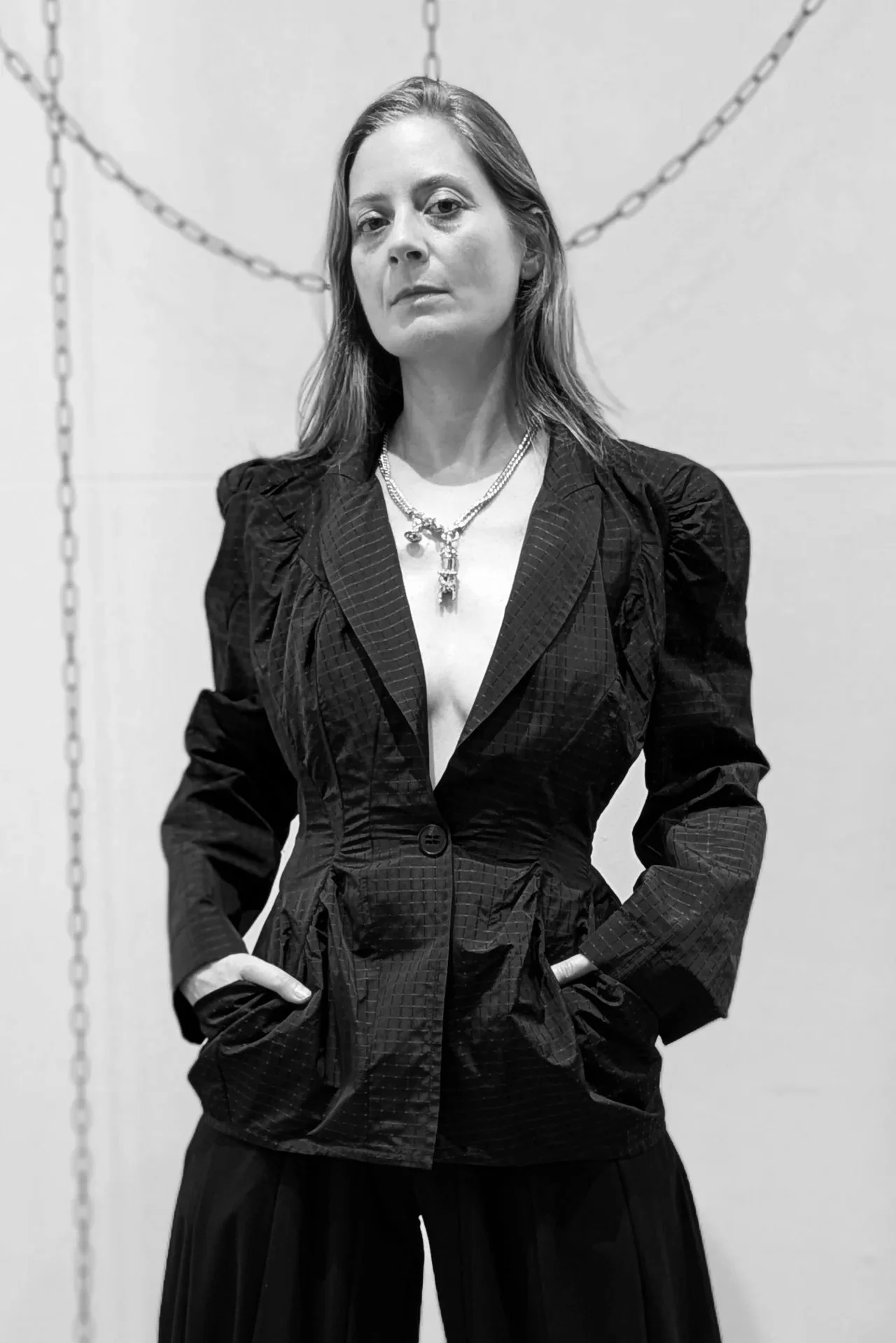 Black-and-white editorial portrait of an artist in a sculptural black jacket, standing against a wall with hanging chains.