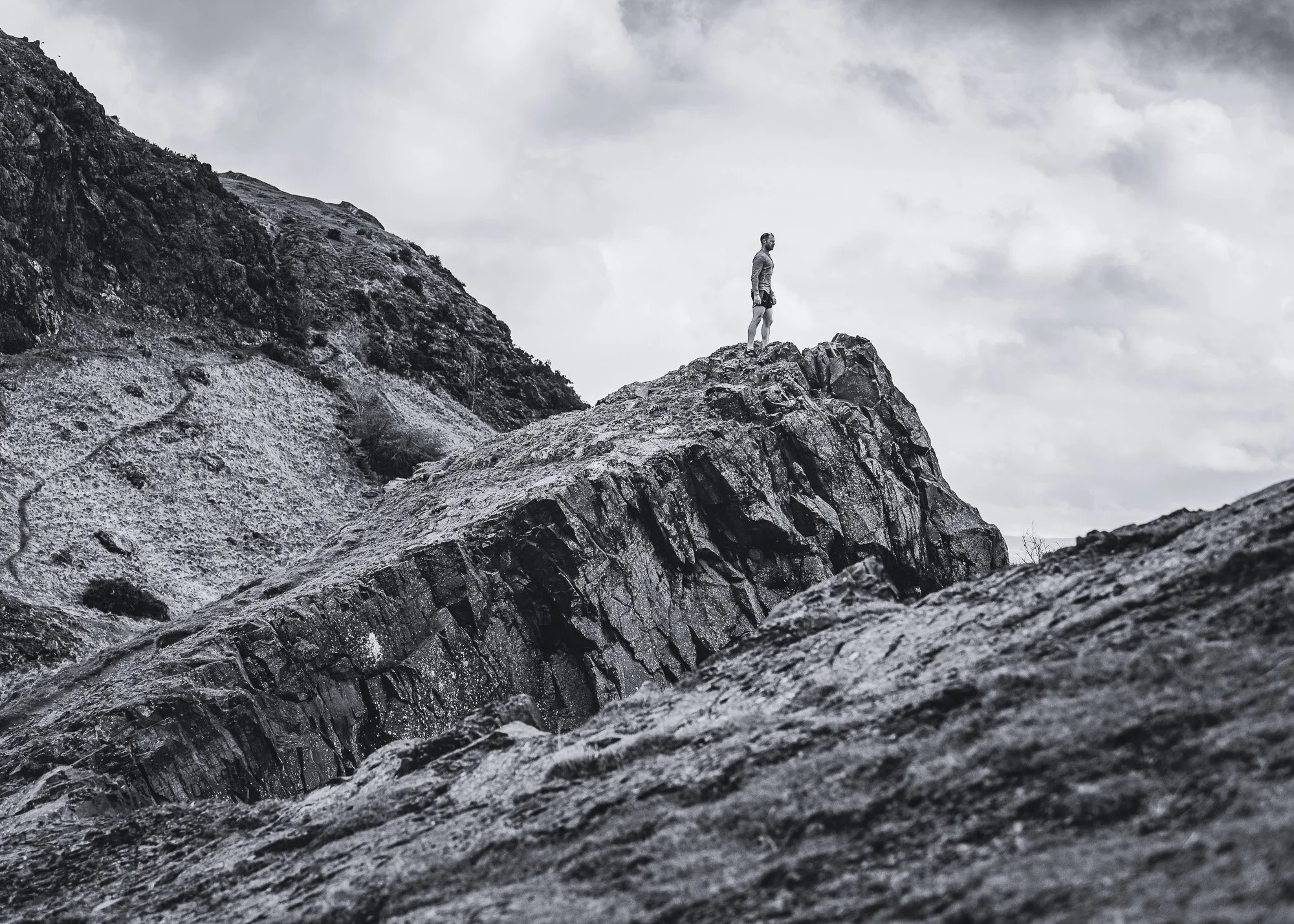 Personal Trainer, Gregor Ridley is standing on a rocky mountain ledge, looking out over a rugged landscape with steep rocky slopes under a cloudy sky. Edinburgh.