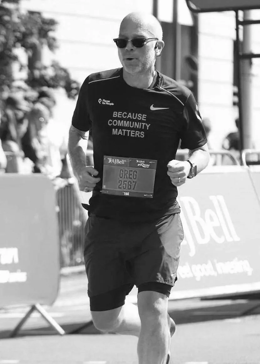 A man running a marathon wearing sunglasses, a sports shirt that says "Because Community Matters," and a bib with the name "Greg" and number "2567."
