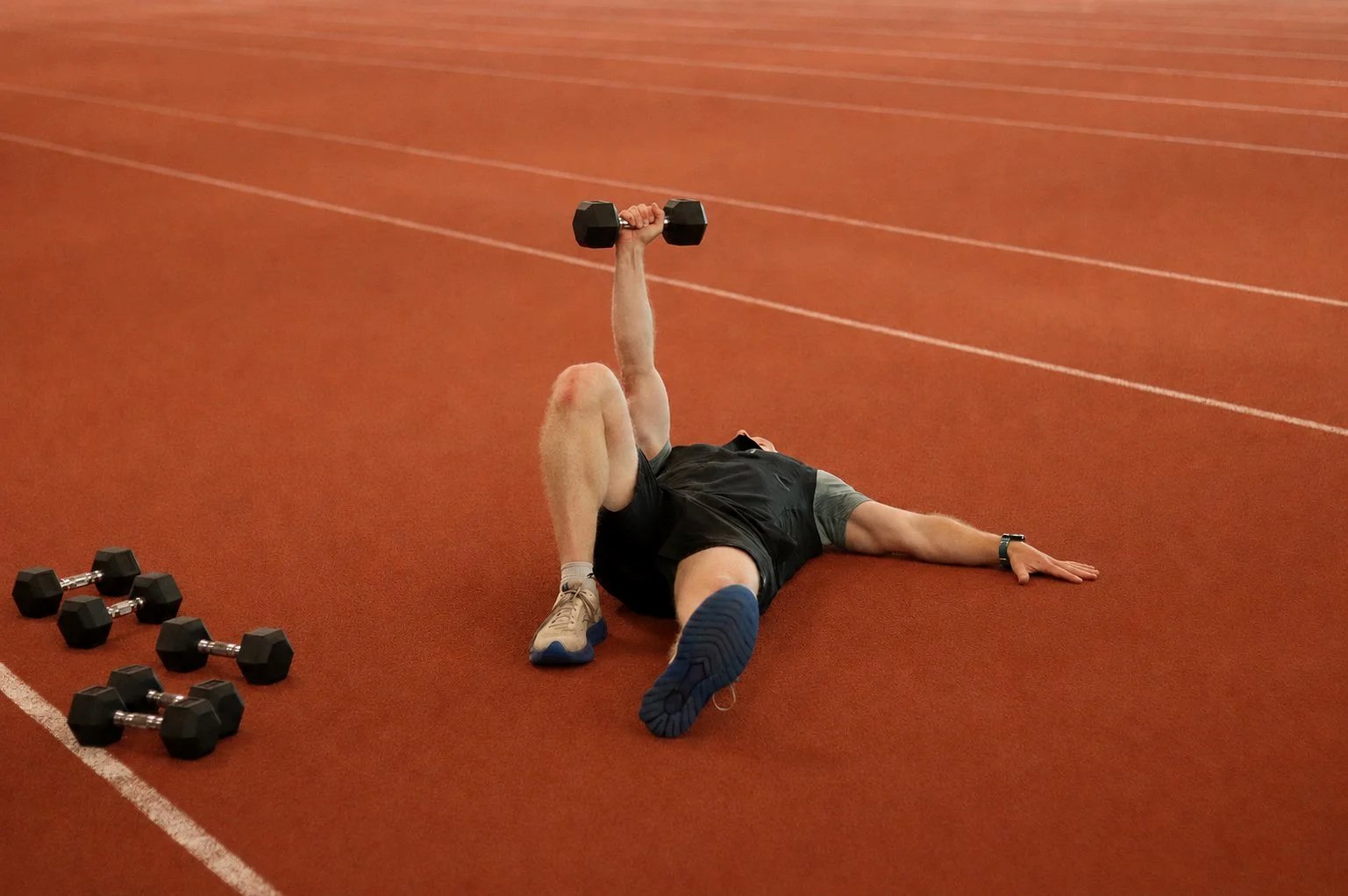 An experienced athlete lying on the running track after completing a workout, holding a dumbbell in one hand. Several dumbbells are on the ground nearby.