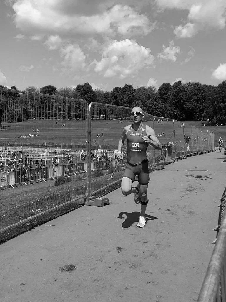 A male athlete wearing sunglasses, a sleeveless sports top with 'GBR' and 'accenture' printed on it, and shorts, running on a track surrounded by a fence in an outdoor park during daytime.