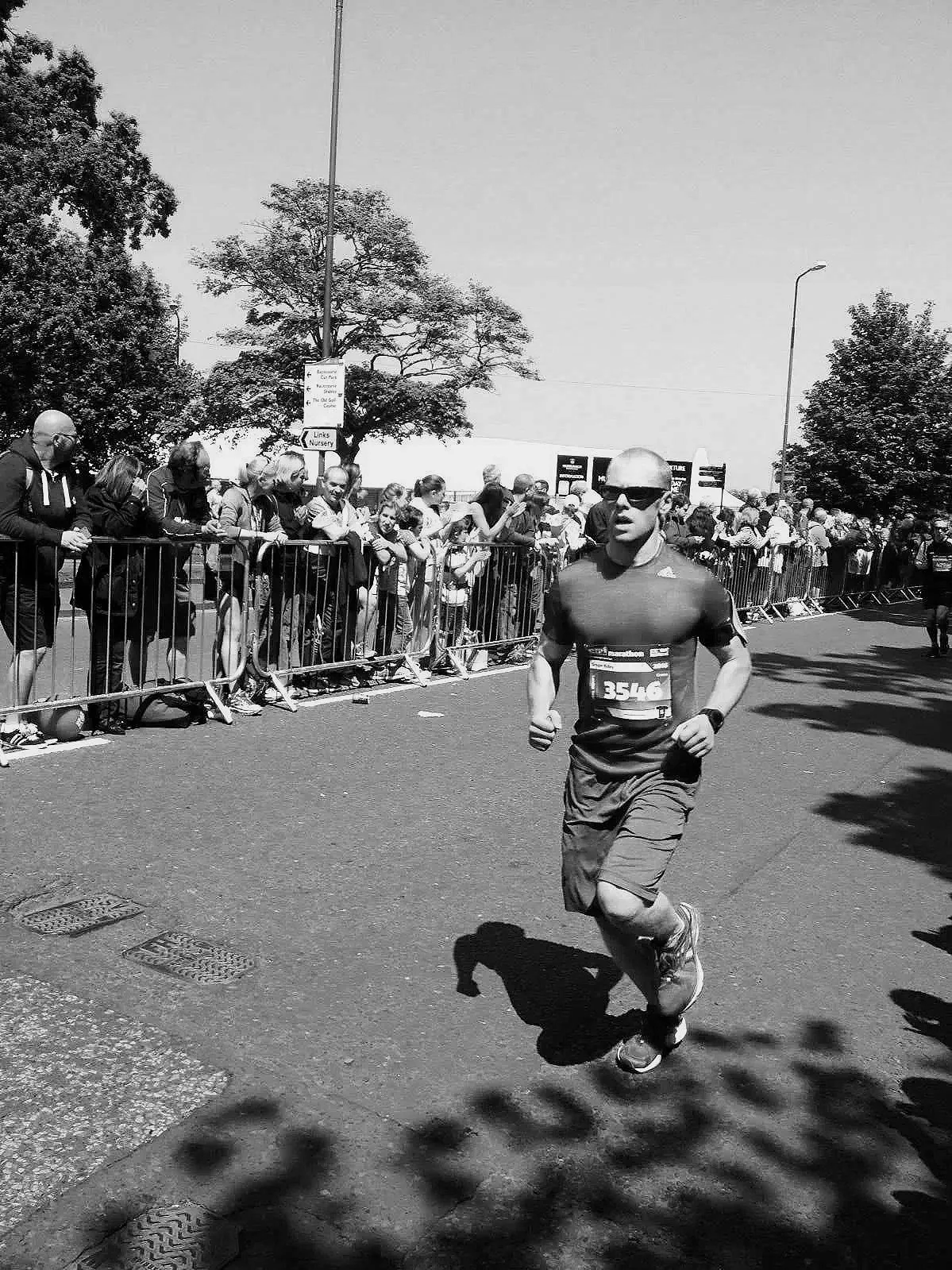 A male runner in athletic clothing and sunglasses participating in a race, with a crowd of spectators behind a barrier watching along the roadside.