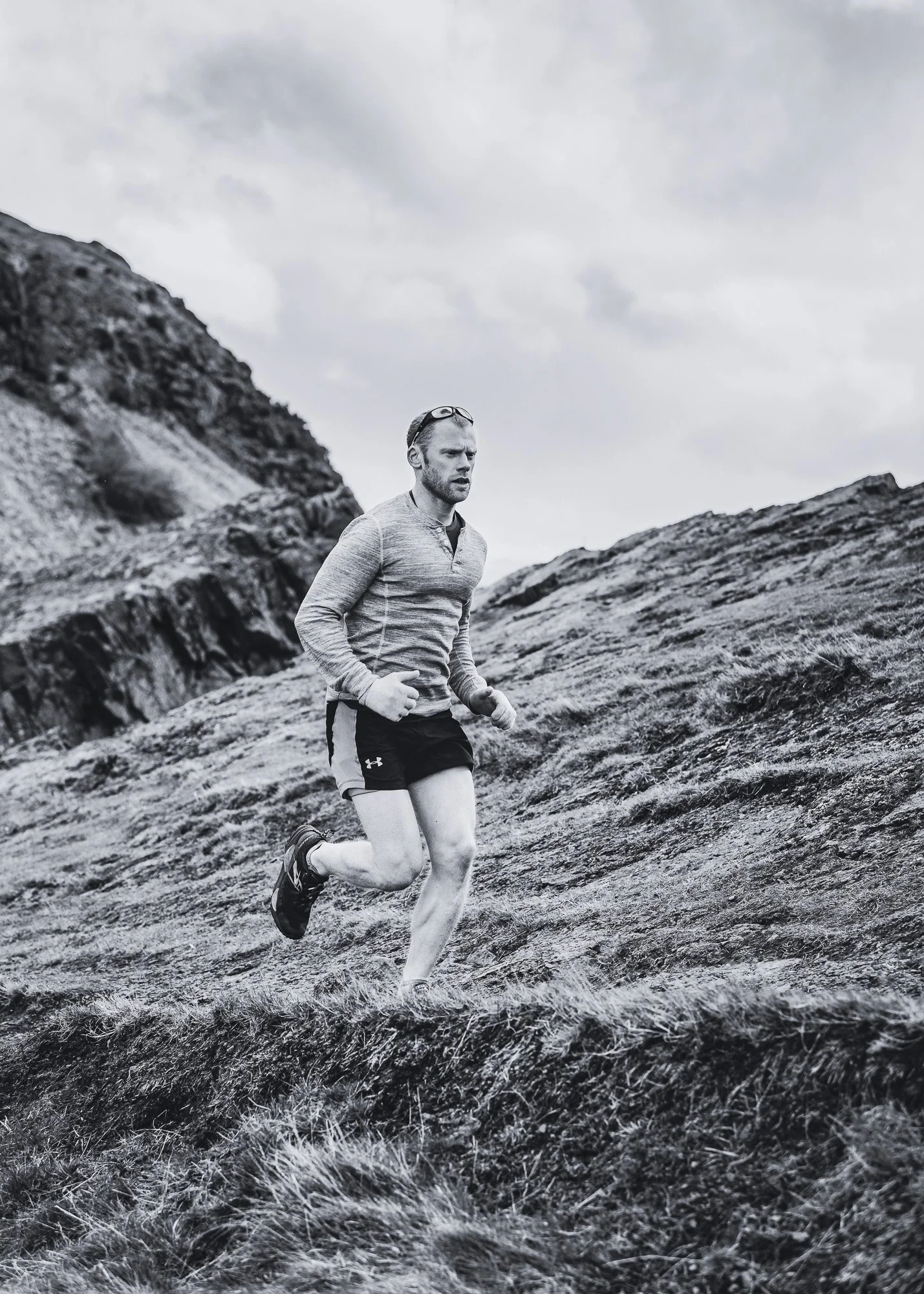 Personal Trainer, Gregor Ridley is running on a rocky, grassy outdoor trail with Arthur's Seat and a cloudy sky in the background.
