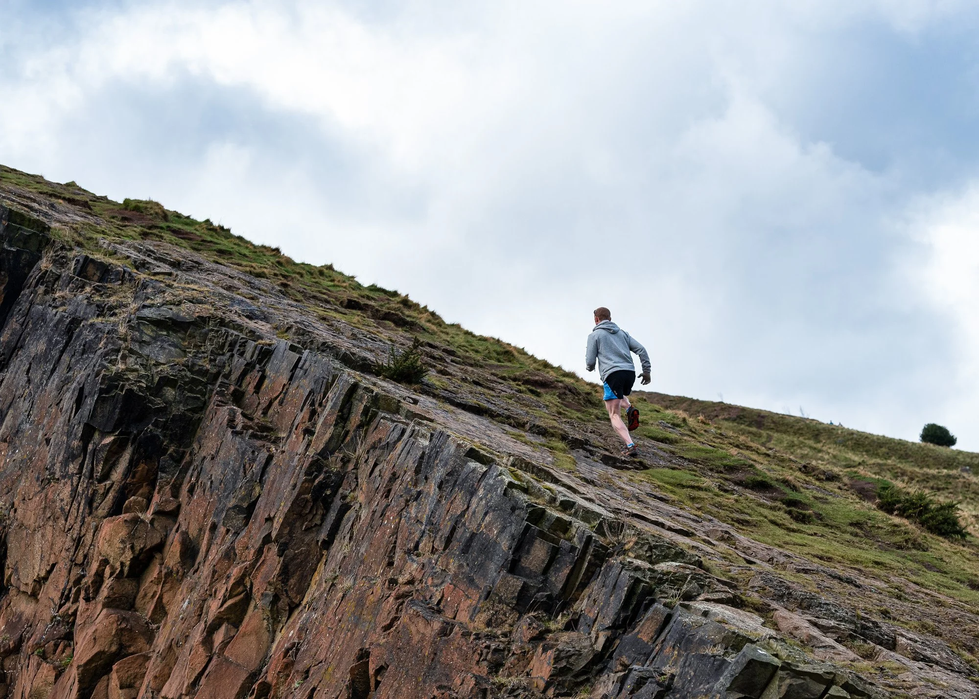 Personal Trainer, Gregor Ridley is running uphill on a rocky and grassy hillside under a cloudy sky in Edinburgh.