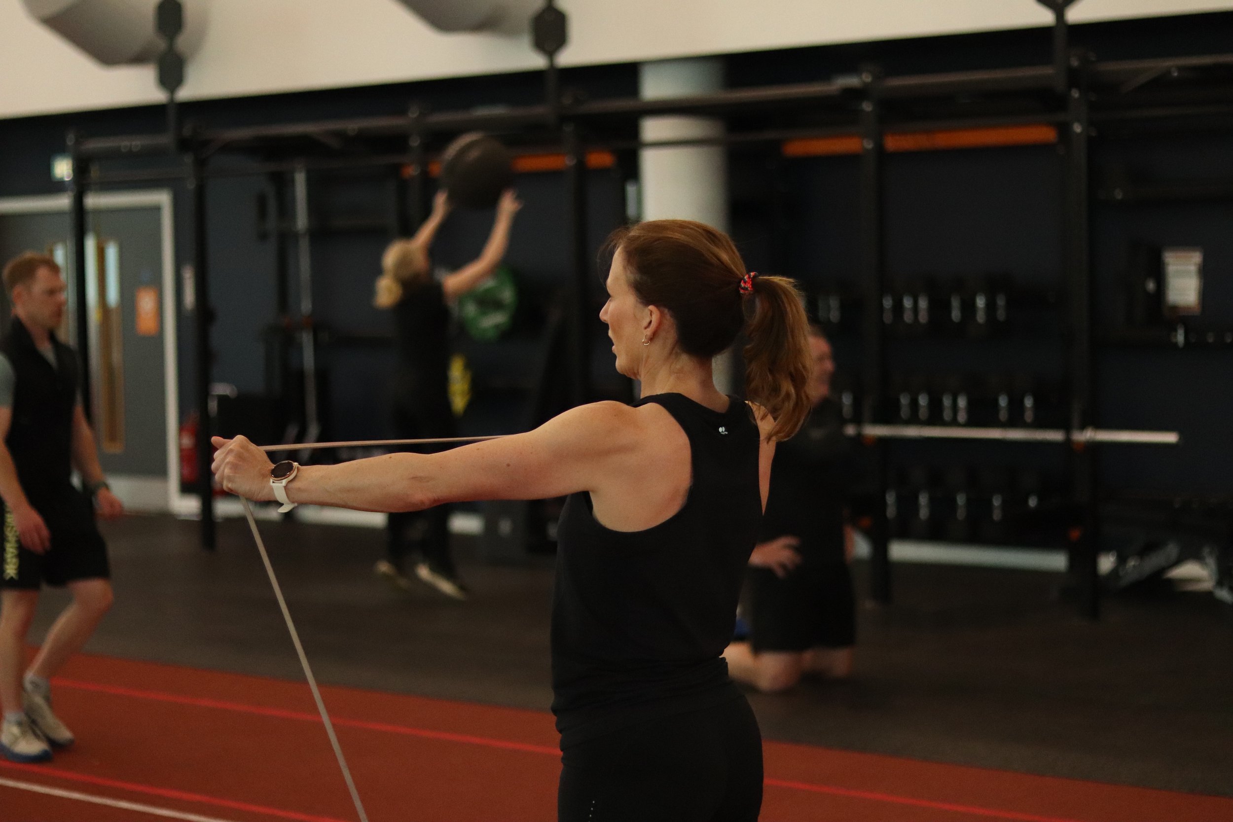 A woman in black workout clothes participating in a fitness coaching session with Gregor Ridley, holding a resistance band, with other people and fitness equipment in the background.
