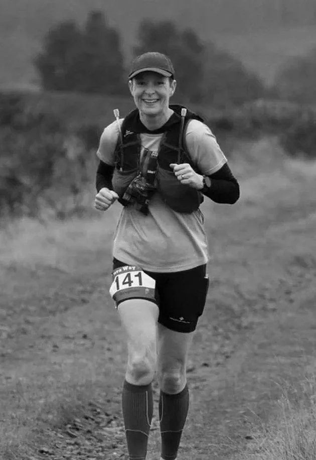 A woman running outdoors on a trail, wearing sports gear including a cap, hydration pack, and athletic clothing, smiling at the camera.