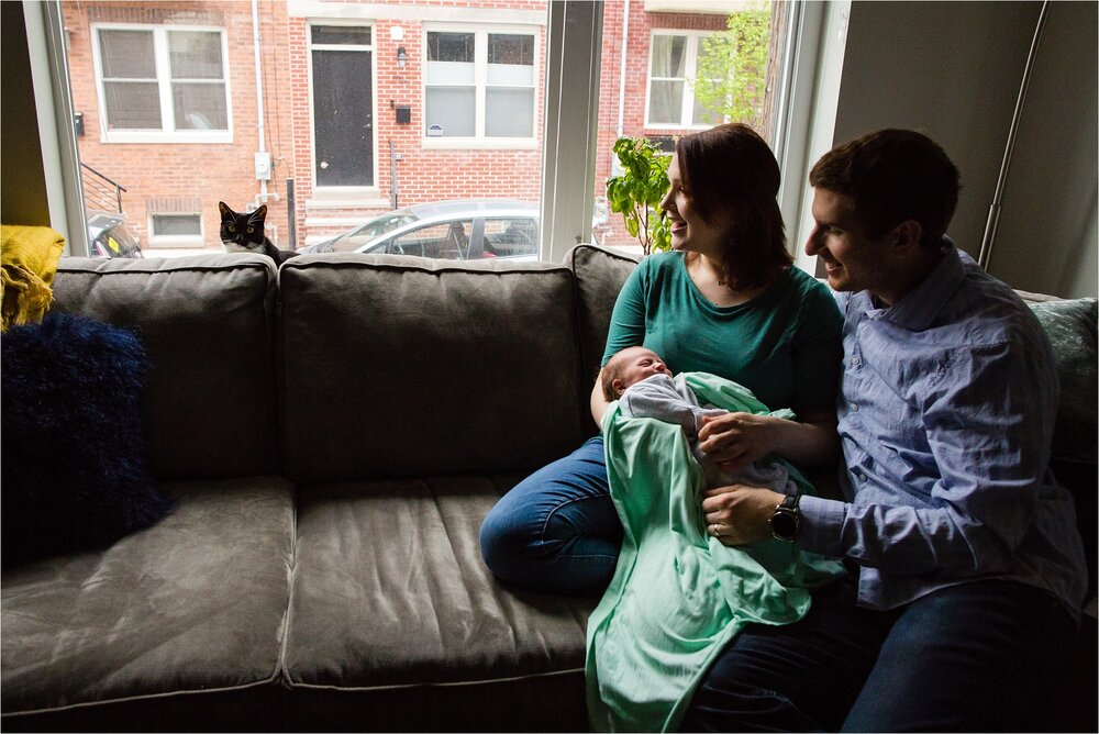 New mom and dad hold baby boy in front of big window, getting ready to breastfeed, and laugh at family cat peeking up from behind the couch, Philadelphia Newborn Photographer