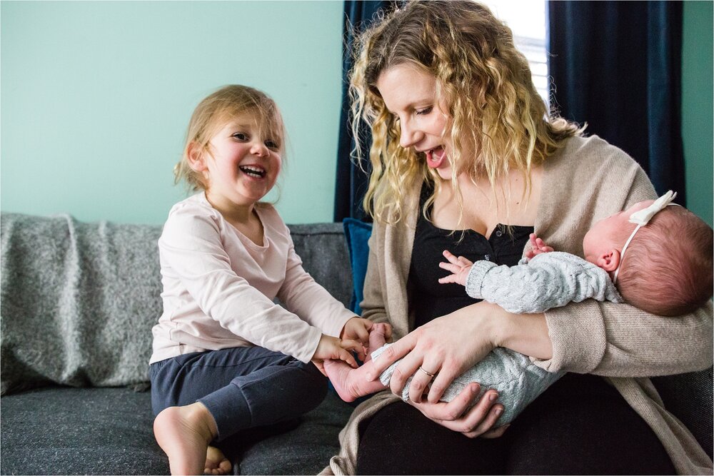 Toddler girl laughs and tickles baby sister's toes while mom laughs and holds her, Philadelphia Newborn Photographer
