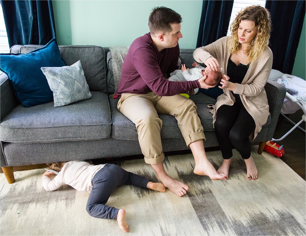 Toddler daughter crawls under the couch while mom and dad tend to baby sister, Philadelphia Newborn Photography