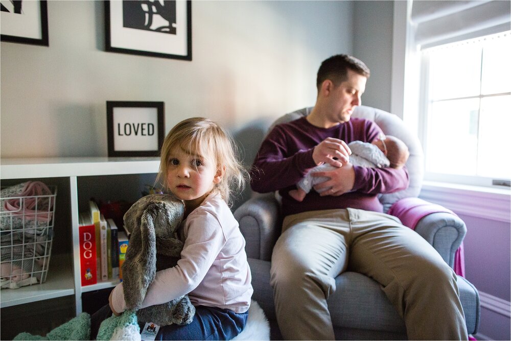 Toddler girl sits on ottoman holds stuffed bunny while dad holds baby sister and sign that says loved in the background, Philadelphia Family Photographer