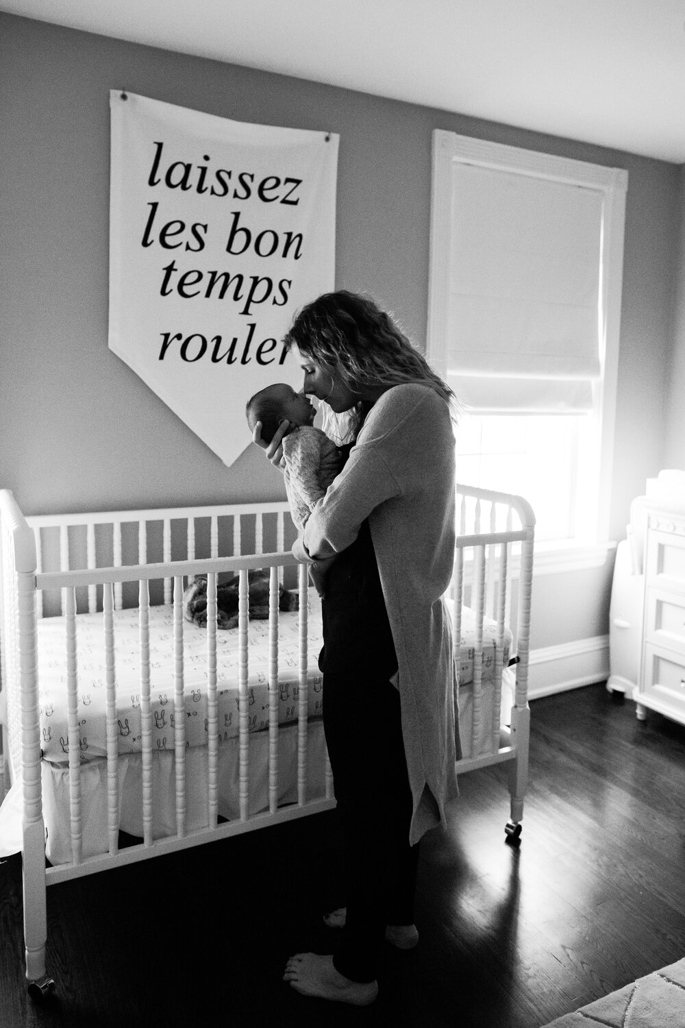 Mother holds new baby in her nursery, in front of the crib and a sign on the wall that reads "let the good times roll" in French, black and white, Philadelphia newborn photography
