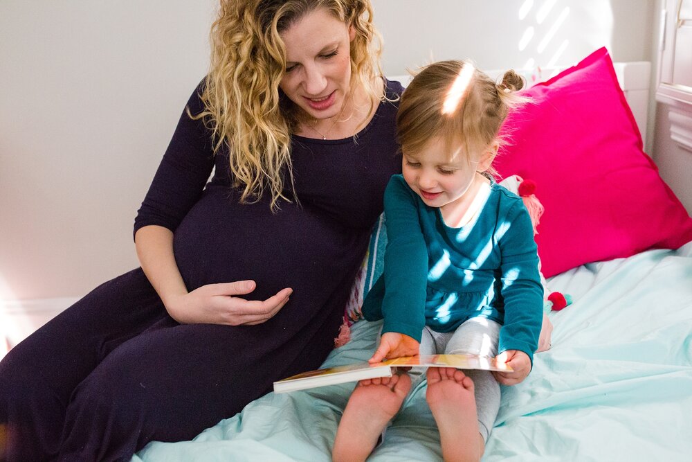 Pregnant mom reads sits on bed with toddler daughter reading a book, hand on her pregnant  belly, dappled window light, Philadelphia maternity photographer