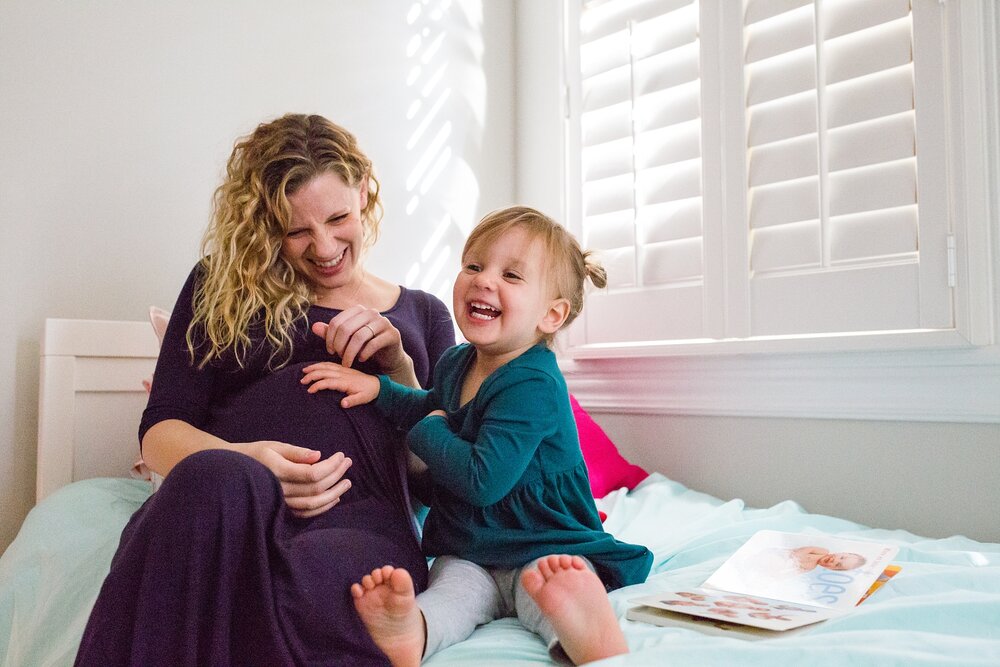 Toddler girl sits on bed with her pregnant mommy and tickles her baby belly, Philadelphia Maternity Photography