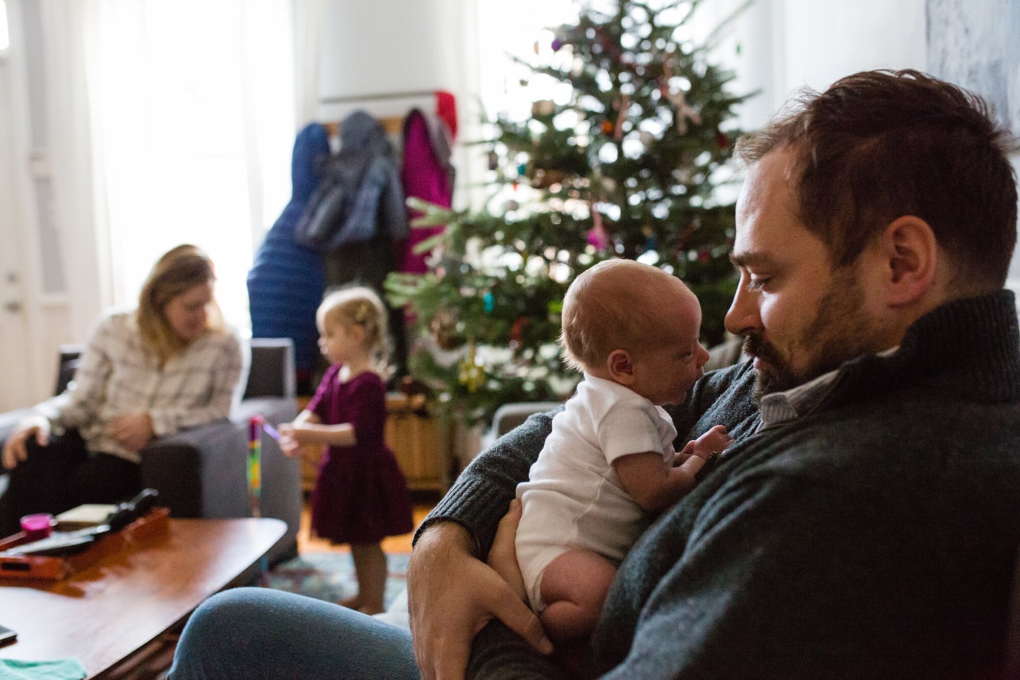 Dad holds baby boy on his chest and looks down at him while toddler and mom play in the background by the Christmas tree, Philadelphia newborn photography