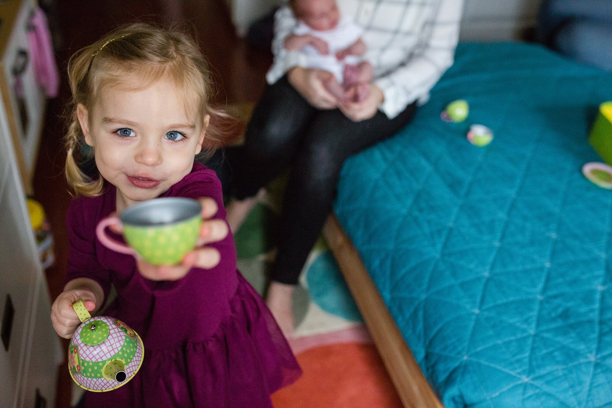 Little girl offers a cup of imaginary tea looking right at the camera, baby brother in the background, Philadelphia newborn photography