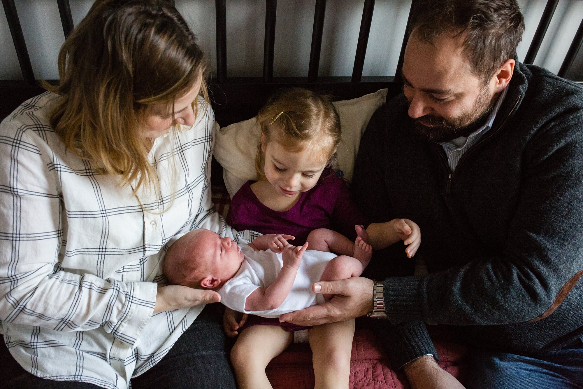 Big sister holds baby brother sitting on the bed with mom and dad, Philadelphia family photographer