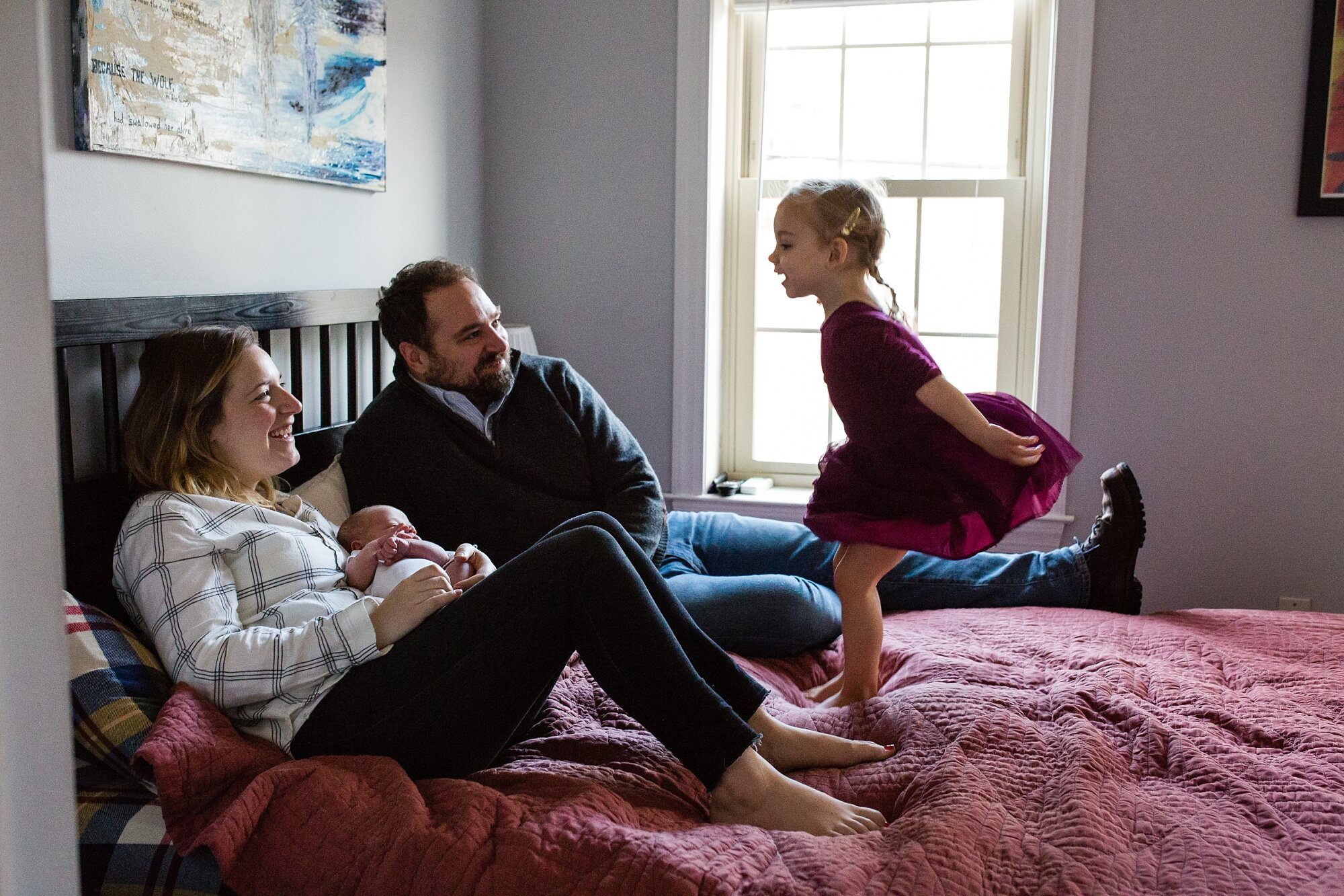 Toddler girl looks coyly at mom and dad holding baby brother, jumps on the bed and laughs, Philadelphia family photographer