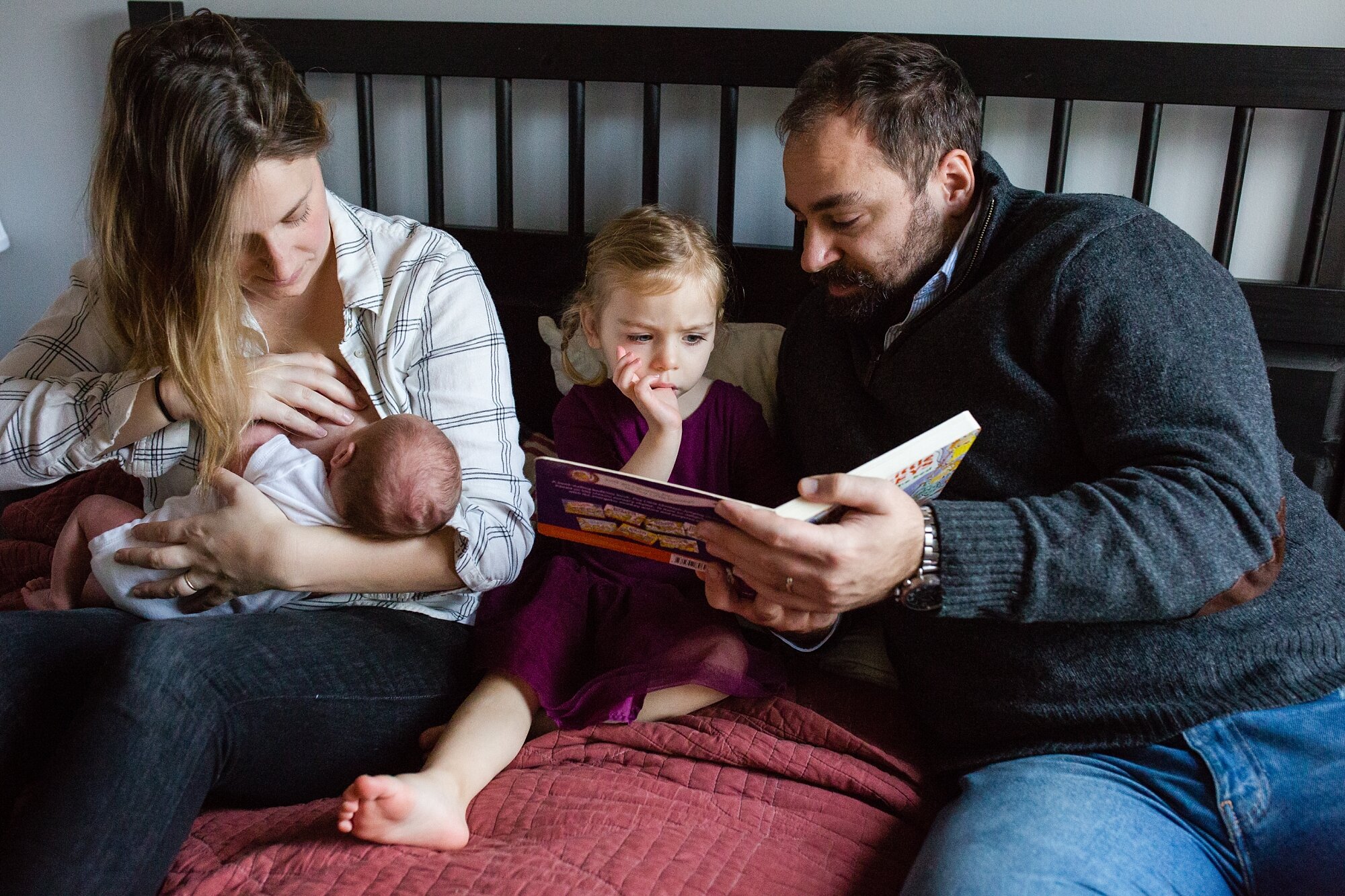 Big sister reads a book with dad while mom breastfeeds baby brother on the bed, Philadelphia family photographer