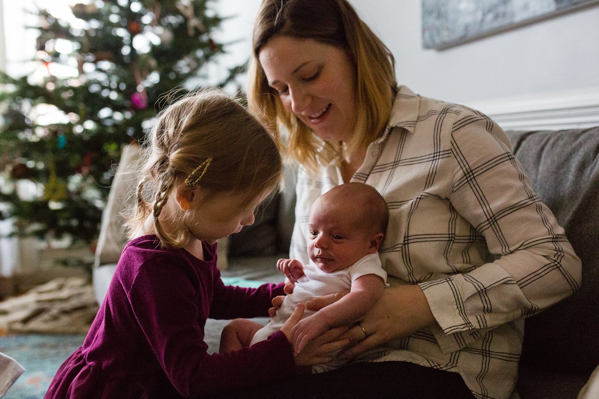 Big sister sibling comes over to say hi to baby brother sitting in mom's lap, Christmas tree in the background, Philadelphia newborn photography