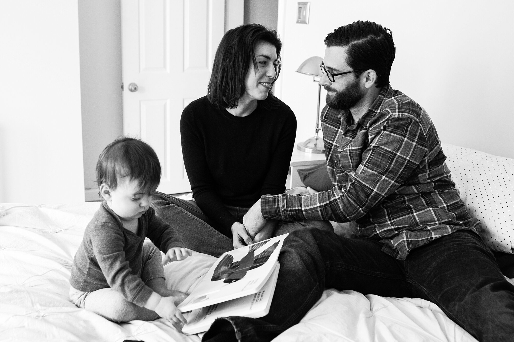 Toddler girl reads her favorite book on the bed while mom and dad steal a glance at each other, and mom scoops daughter up for a hug and kiss, Philadelphia Natural Light Photography