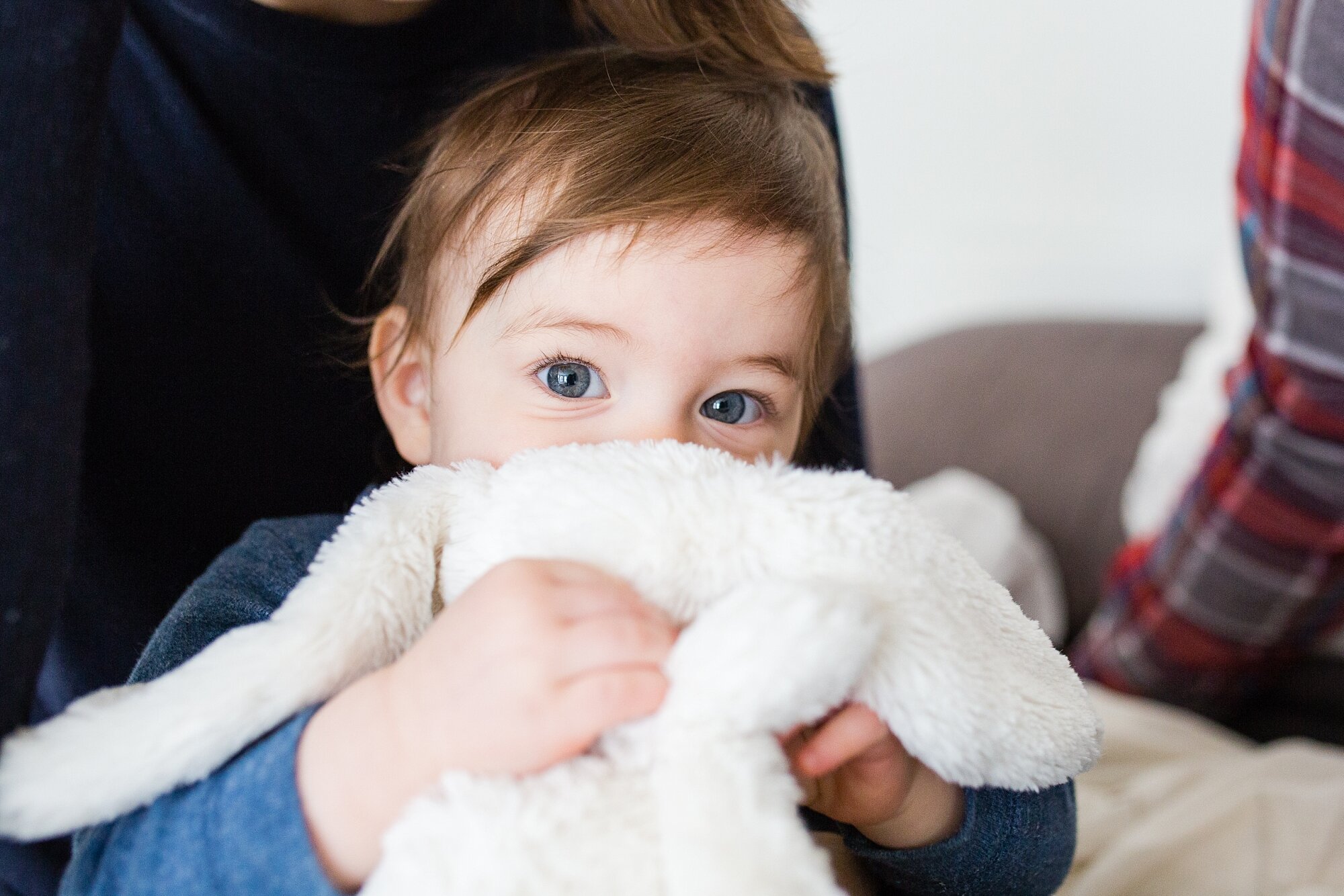 One year old girl with big gorgeous blue eyes peaks over her white stuffed bunny and smiles at the camera, Philadelphia Natural Light Photographer