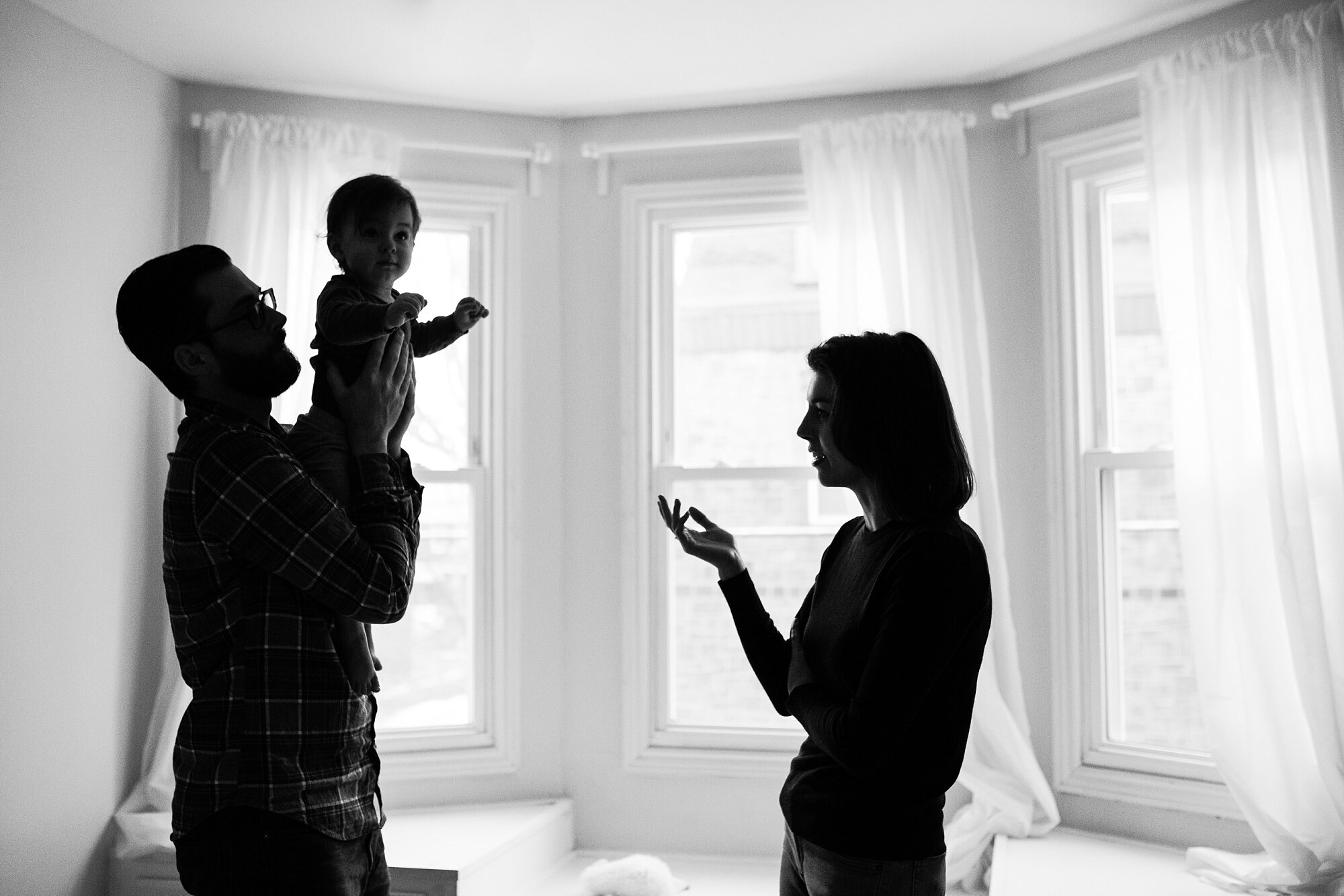 Black and white portrait of a family of three, silhouette of dad holding toddler while mom is talking, Philadelphia family photographer