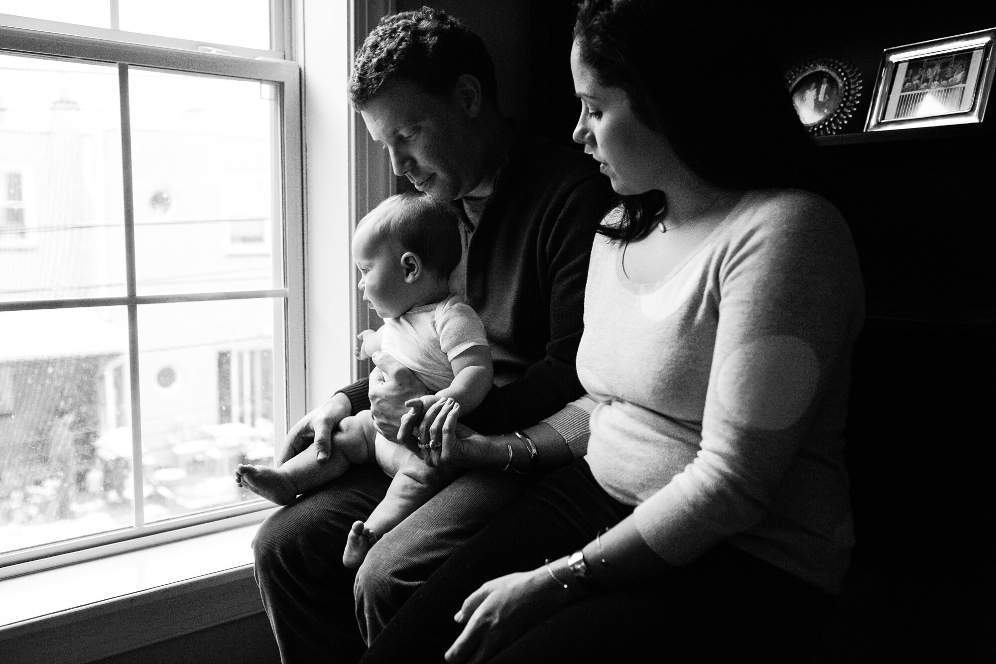 Family of three sits by the window watching snow fall, black and white portrait, Philadelphia Baby Photographer