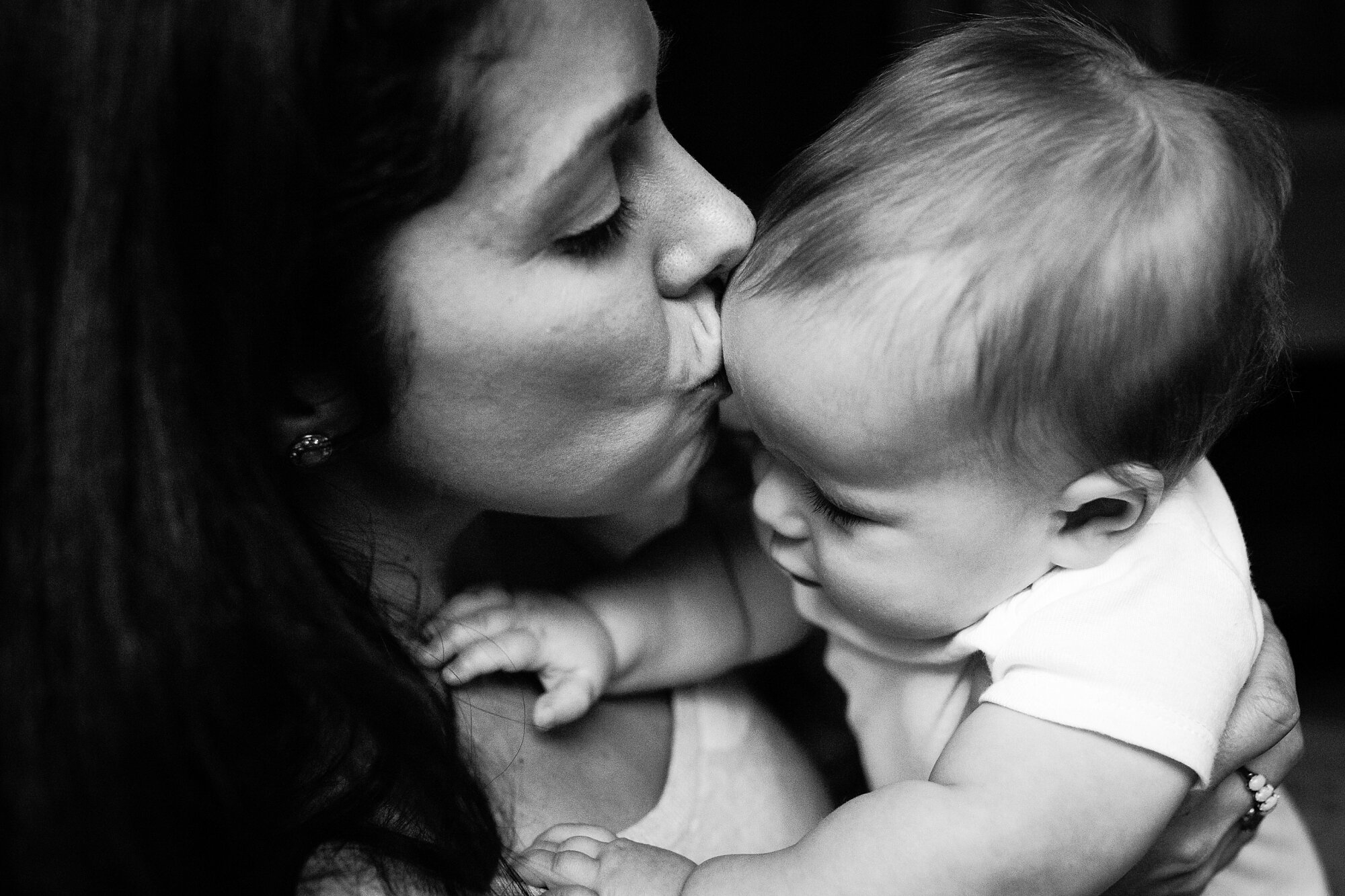 Mom kisses eight month old son's forehead, baby reaches for his mommy, home on a snow day, black and white portrait, Philadelphia Family Photography