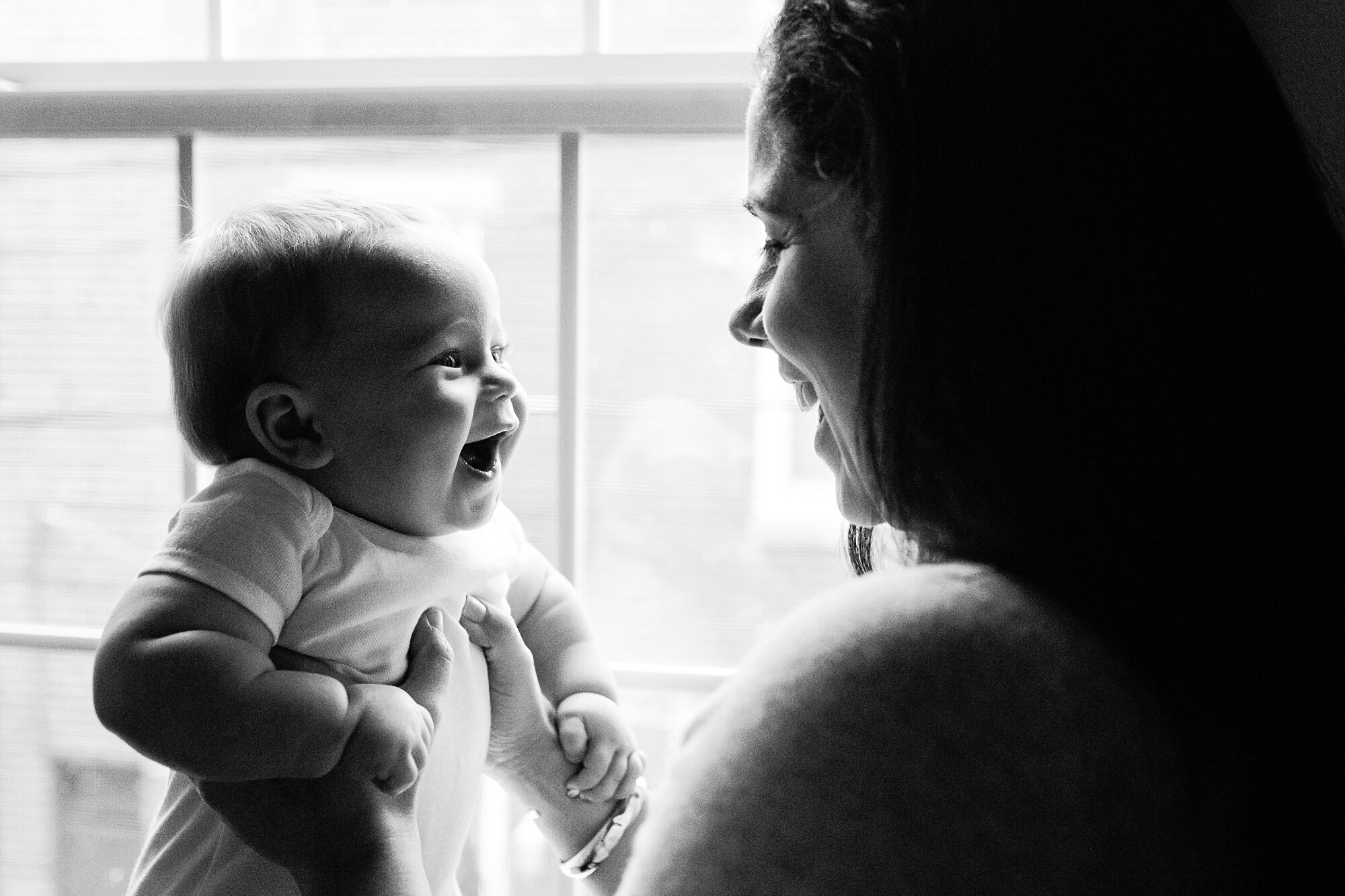 Mama and baby son smile and laugh together, sitting by the window watching the snow, black and white portrait of motherhood, Philadelphia Family Photographer