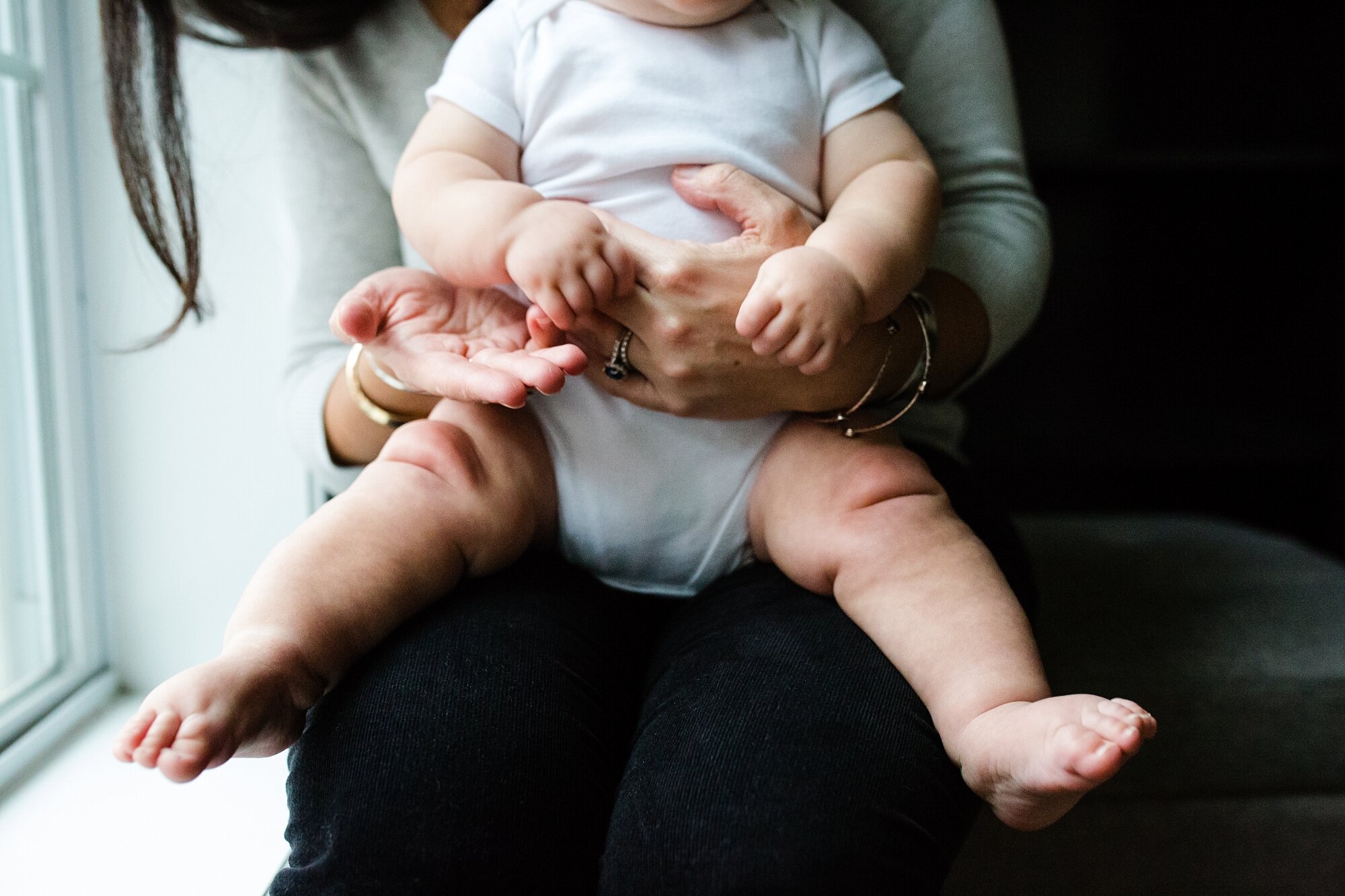 Baby with the chubbiest legs sits on mom's lap in front of the window, home on a snow day, Philadelphia Family Photographer