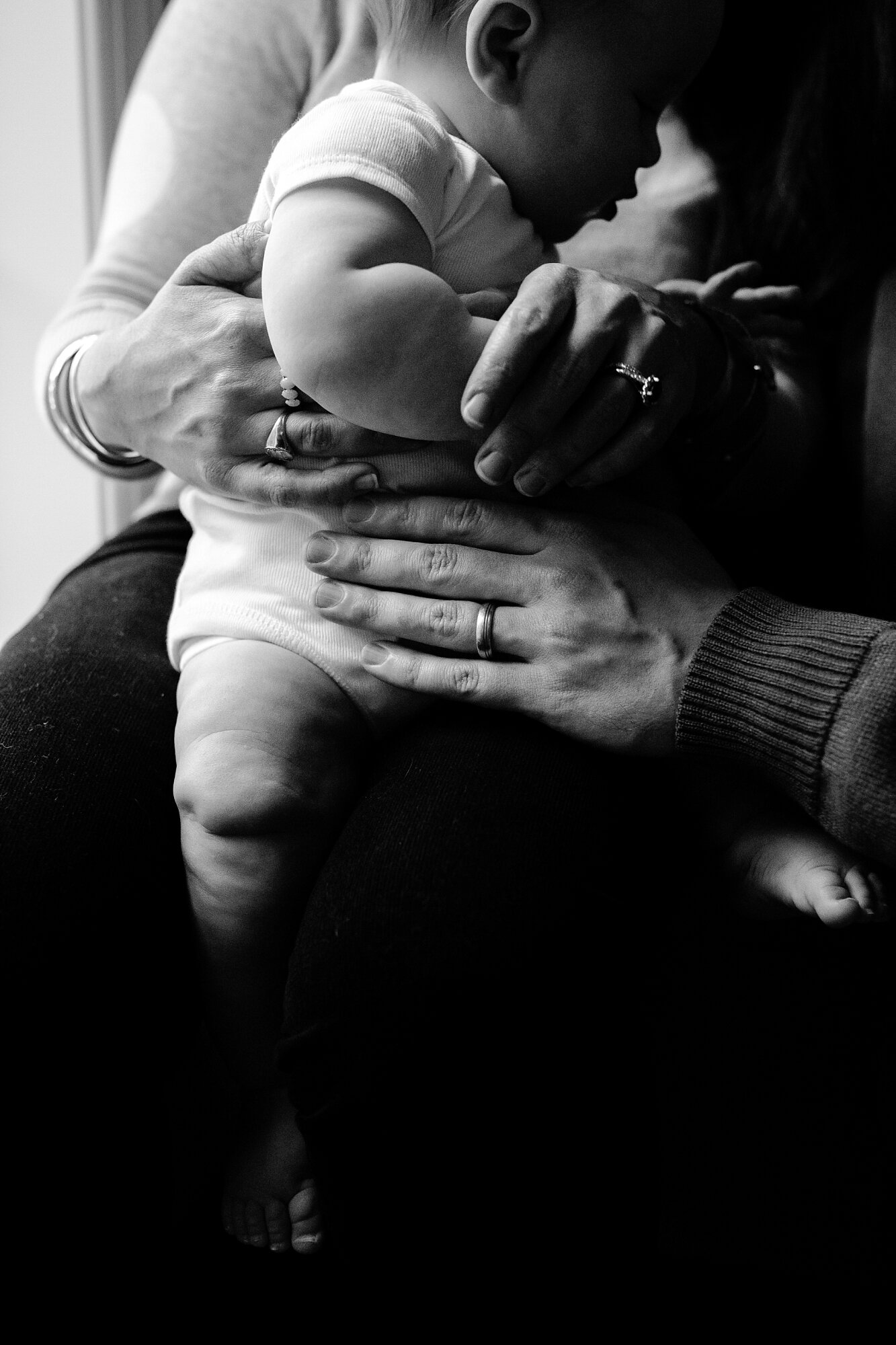 Black and white portrait of mom, dad, and baby in front of a window, parents' hands on son whose face is in silhouette, Philadelphia family photographer