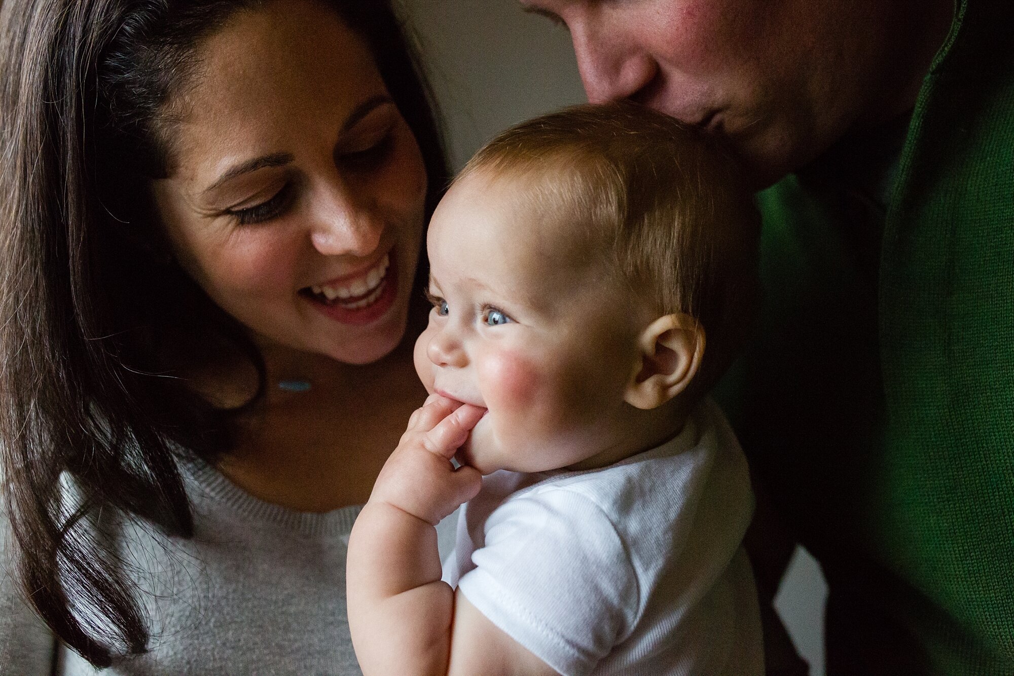 Mommy daddy and baby sit by the window with snow falling outside, laughing and kissing, Philadelphia Family Photographer