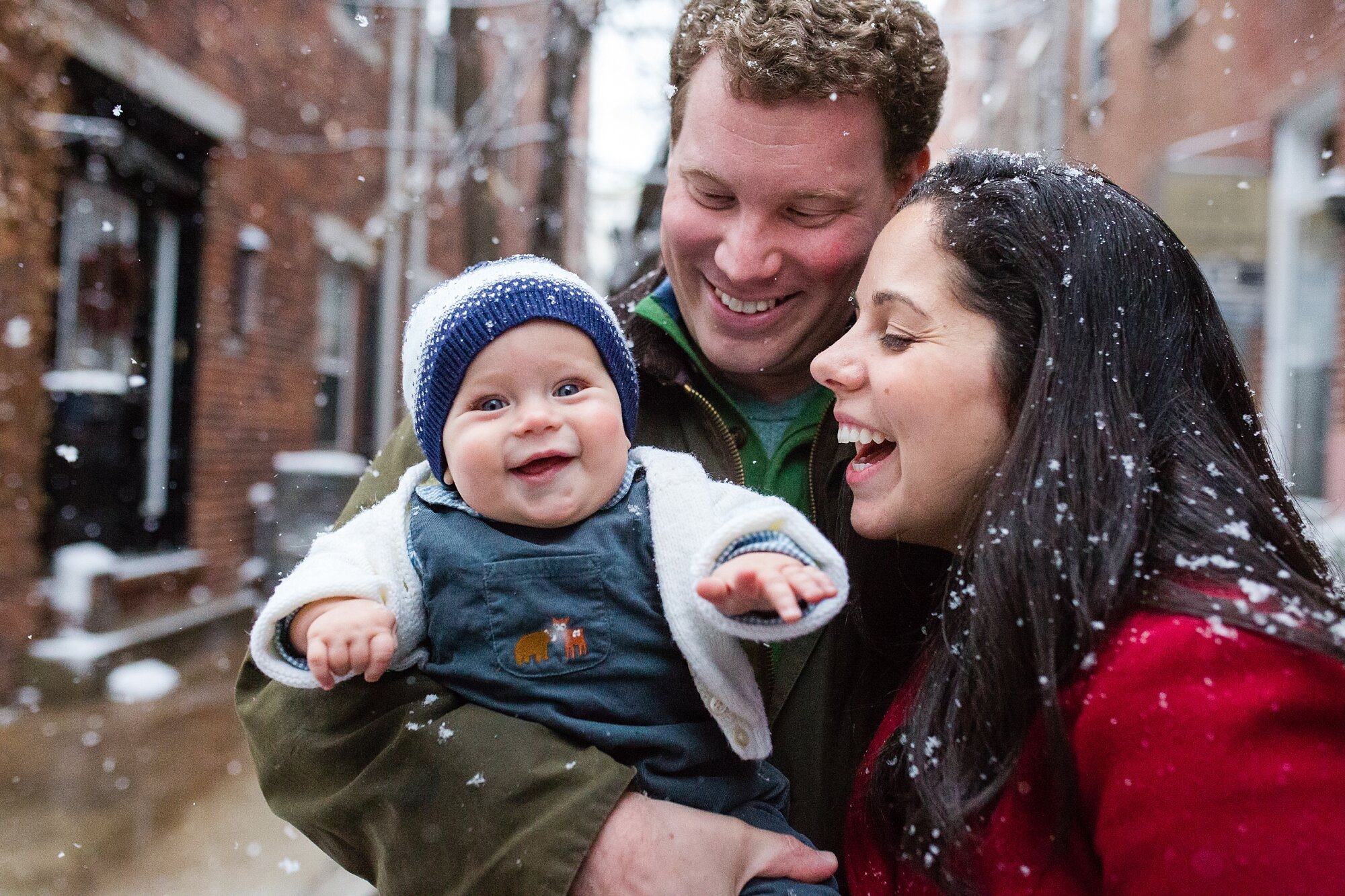 Family of three take 8 month old son outside to experience his first snow fall, baby laughs and smiles, mom wearing red coat, Philadelphia Family Photographer