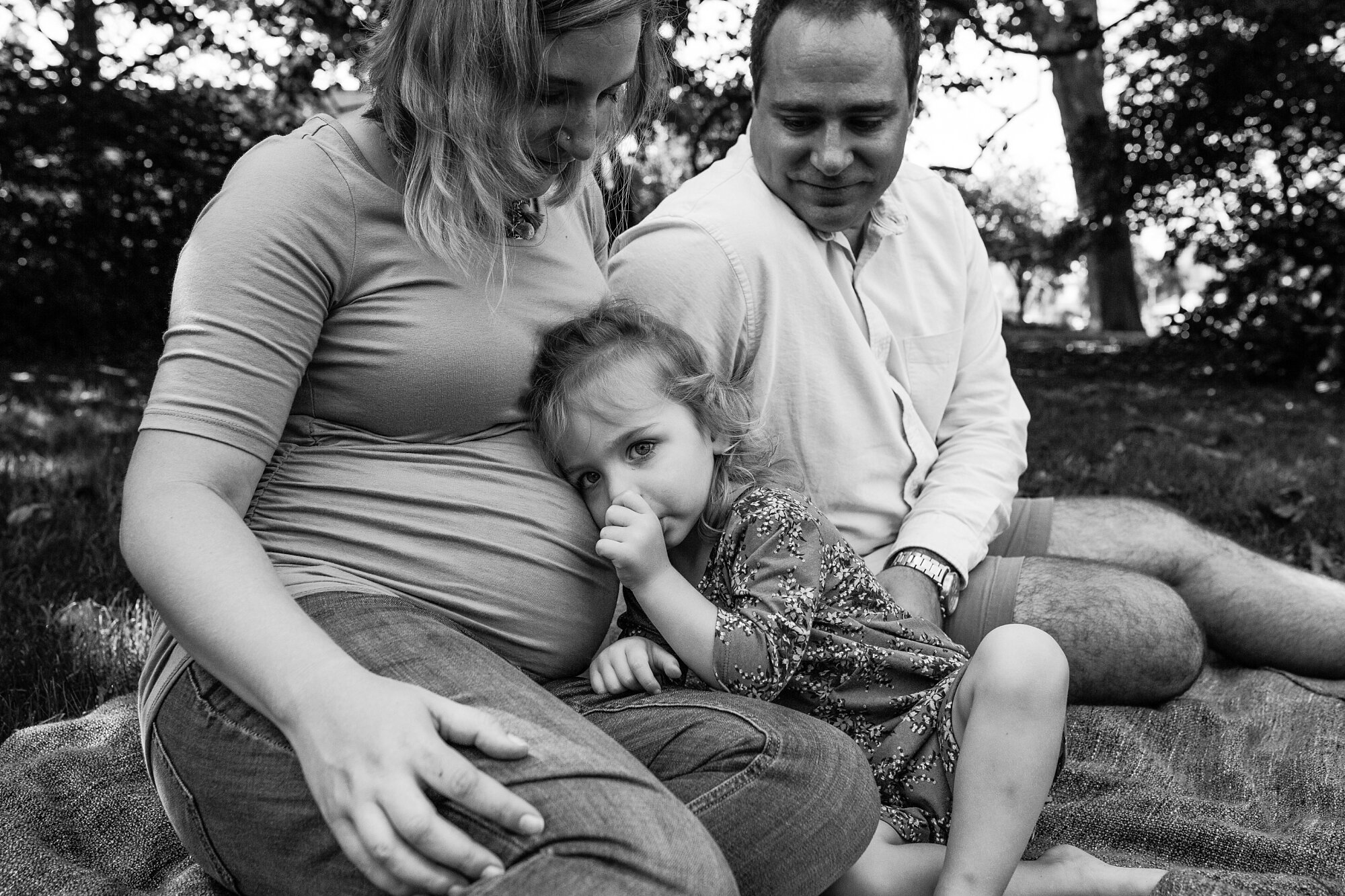 Family of three poses on a blanket in the grass, toddler rests her head on pregnant mom's belly and sucks her thumb at the Azalea Gardens, Fairmount, Philadelphia Family Photographer