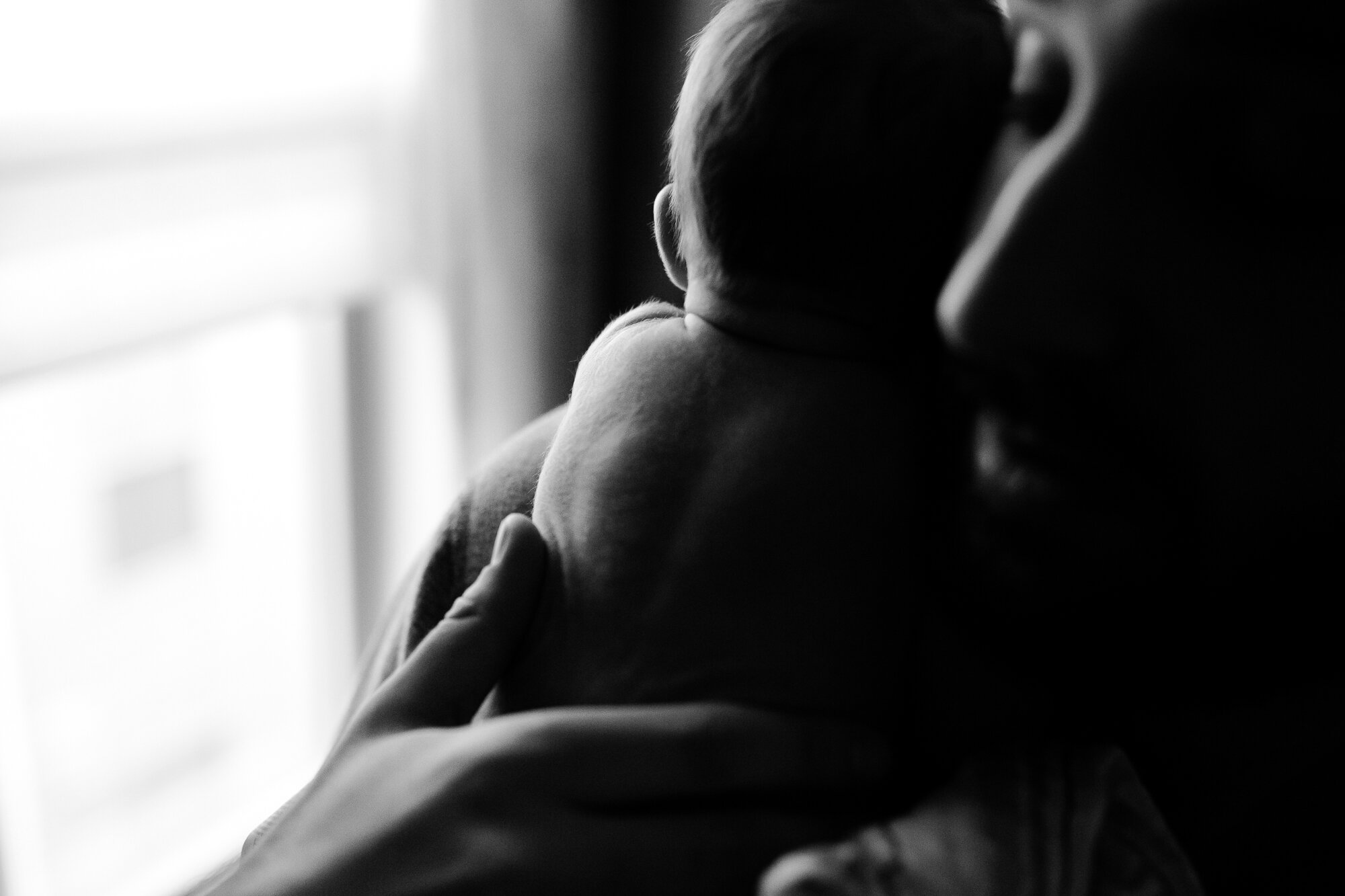 Black and white image with rich shadows and light shining on the tiny hair on baby's back as he rests on his father's shoulder, Philadelphia Newborn Photography