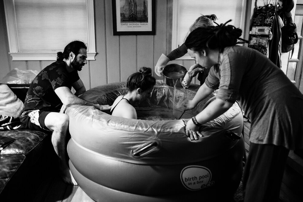 Black and white portrait of a home birth, mom labors in a birth tub, husband gives her support and comfort, midwives pour hot water and check the temperature for her, birth photographer Philadelphia