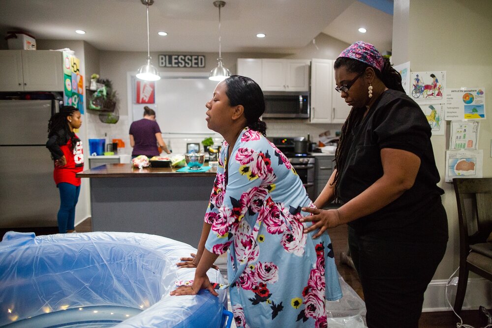 Black mother laughs while in labor, receives hip pressure from her midwife while her doula sets up the birth tub, home birth Photographer Philadelphia