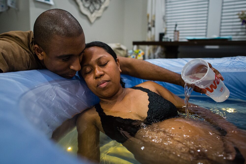 Black dad pours water on his wife's pregnant belly while she labors in the tub, Philadelphia home birth photography
