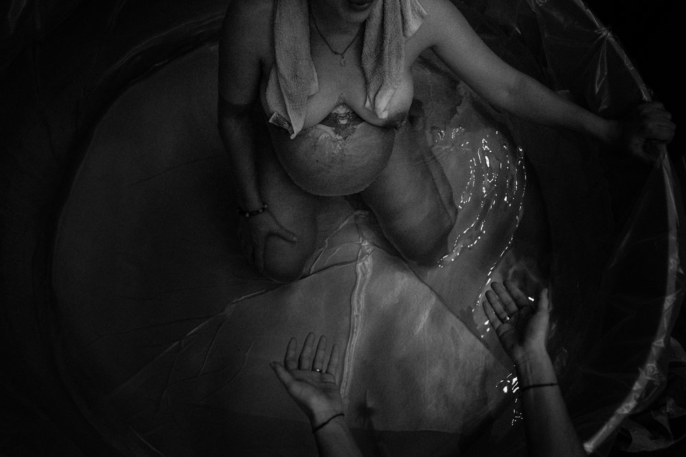 Black and white portrait of a woman in labor in a birth pool, towel around her neck, her doula's hands rest open across from her.