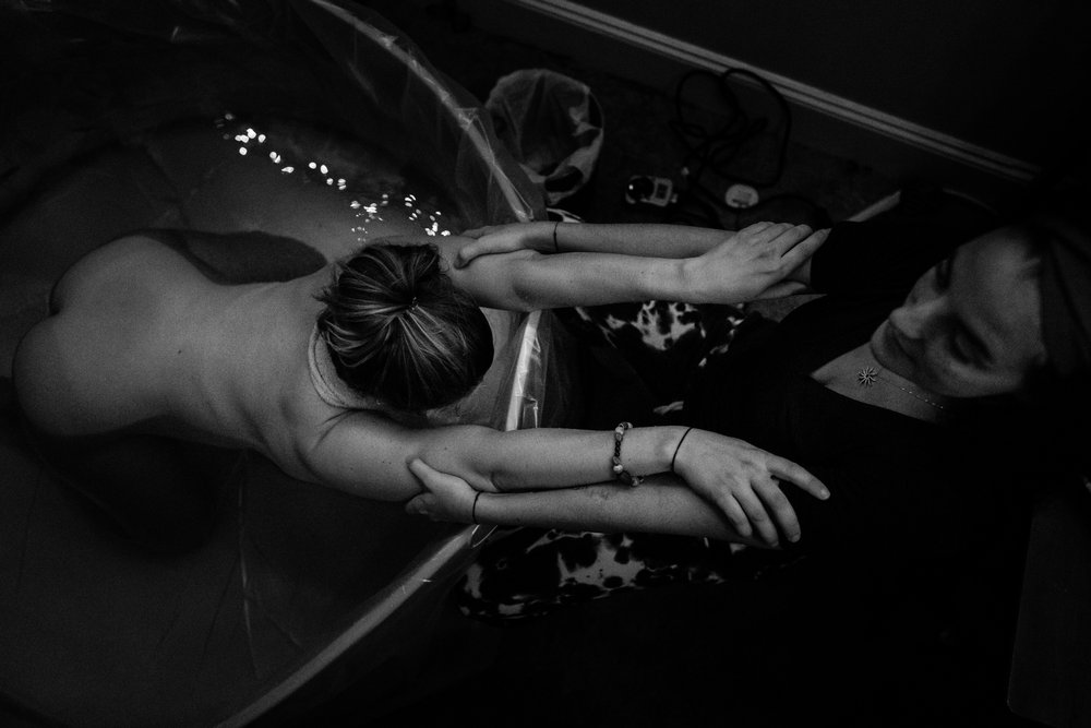 Woman in labor leans over the side of the birth pool and rests in her doula's arms, black and white portrait