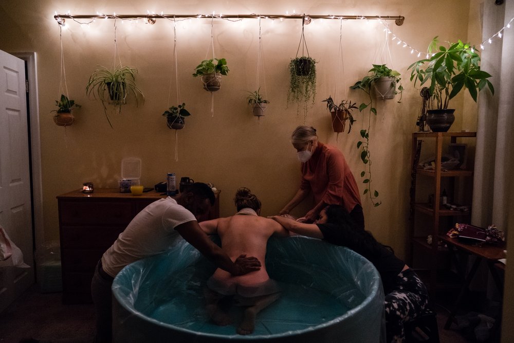 Woman in labor in birth pool gets support from her partner, doula, and midwife, wall of plants and lights in the background