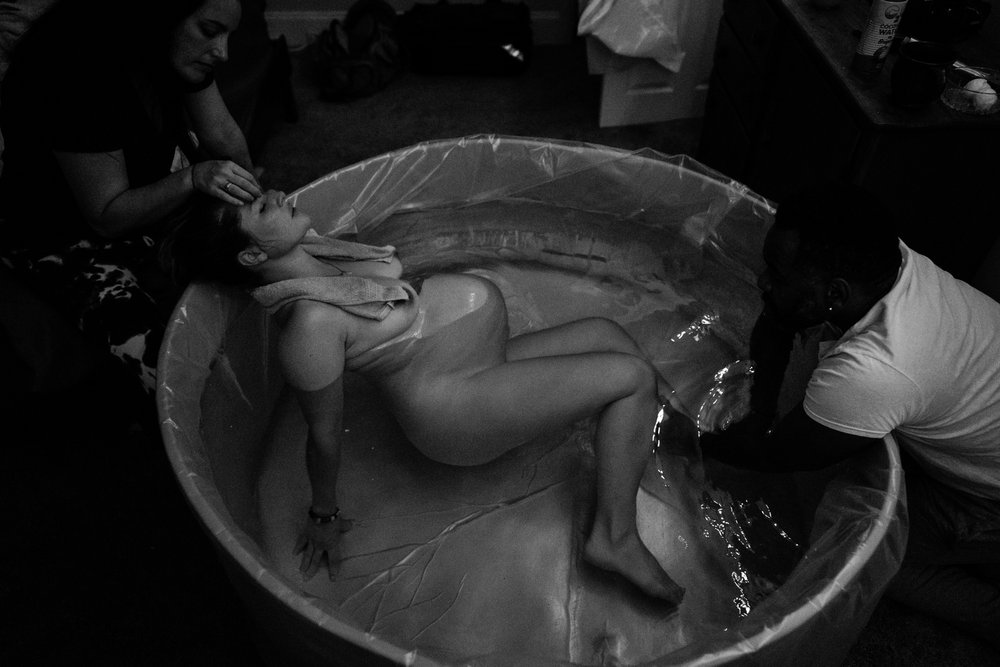 Woman laboring in a birth tub, her belly visibly contracting, her doula rubbing her head, her partner massaging her legs, black and white photo