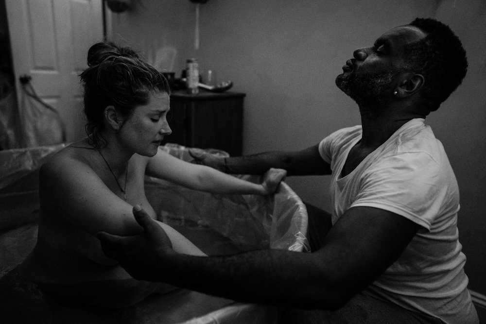 Black father leans back with eyes closed as his partner rests between contractions in birth pool