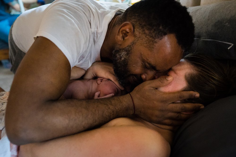 Black father embraces his partner, they kiss, oxytocin flowing, after the birth of their baby snuggled in between them